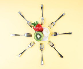 Creative composition in the form of the sun made of forks arranged around a tiny plate with a piece of banana, half of a kiwi and a strawberry. Top refreshing and energy boost fruits snack concept.