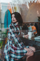 A young girl sits on a chair in a coffee shop and looks at the street through the window of the cafe.