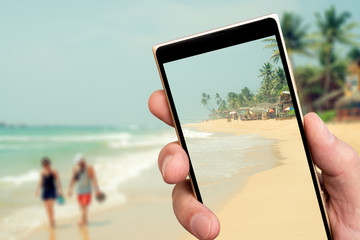 Sandy beach by ocean on smartphone screen. Summer sunny day in tropics. Palm trees on beach.