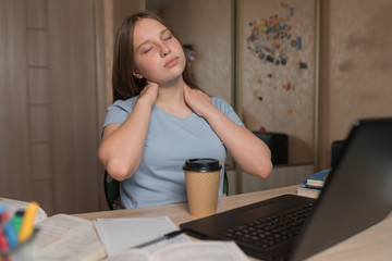 Girl teenager, tired kneads neck muscles, long work and exhausted, fatigue drowsiness, home electronic education laptop Internet. School education preparation college. A cup coffee with tea.