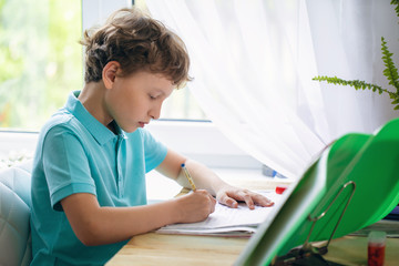 attentive boy writing in notebook while sitting at desk and doing homework.