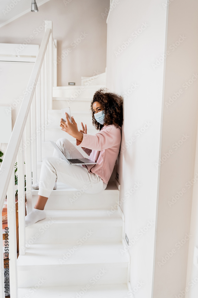 Sticker african american freelancer in medical mask sitting on stairs and showing peace sign while taking selfie near laptop