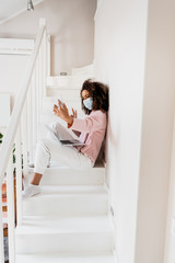 african american freelancer in medical mask sitting on stairs and showing peace sign while taking selfie near laptop