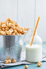 A bucket with a sweet caramel popcorn and a bottle of milk and straws on a worn blue table.