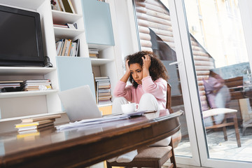 selective focus of tired african american freelancer looking at laptop near documents, books and cup