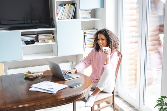 happy african american freelancer holding cup while using laptop at home