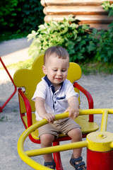 a one-year-old cheerful boy in a t-shirt  sitting on a  children's carousel in the summer