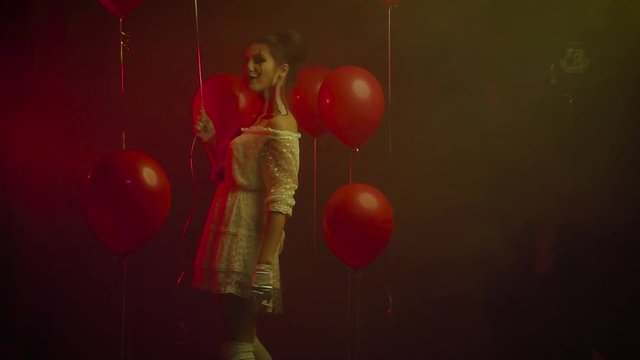 Pennywise breaks into nightmare, evil clown plays with red balloons, lady in white short dress with creative bloody make up, aggressive joker kicks at camera, unusual model plays to advertise show