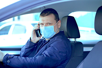 A man in a protective mask is looking at the camera, drives behind the wheel with right-hand drive and receives calls by mobile phone.Virus protection concept, COVID-19