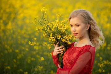 Naklejka premium Portrait of blond teen girl wearing at red dress posing at field with yellow rapeseed flowers. . Happyness and summer time concept
