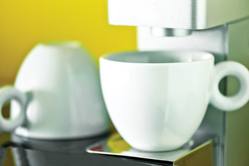 Coffee mugs with coffee machine, espresso, with blurry mug at the background, close up. Several white clean coffee mugs and saucers on a shelf in the pantry.
