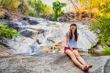 Asian woman travelers are smiling and relaxes sitting on the stone in front of the waterfall at Namtok Kaew Chan., Ratchaburi, Thailand.