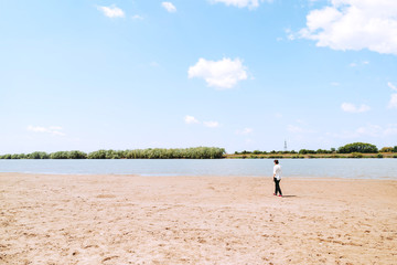 Fototapeta na wymiar A girl in a white shirt walks alone by the river Bank. Small figure walks along the sand