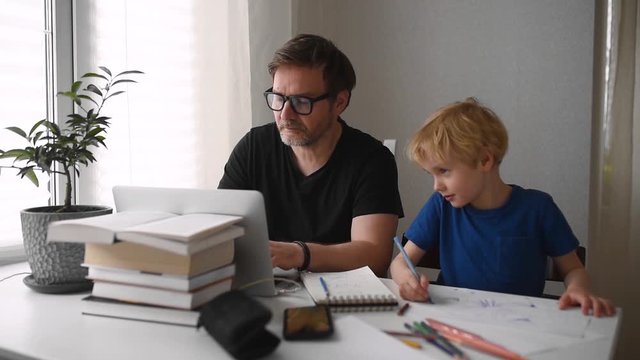 Man working from home with laptop during quarantine. Home office and parenthood at same time. Preschooler child drawing while parent freelancer making job. Difficulties with kids during isolation