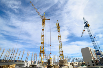 Naklejka premium Photos of high-rise construction cranes and an unfinished house against a blue sky. Photographed on a wide angle lens.