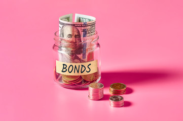 Glass jar with coins and inscription bonds lies on pink background