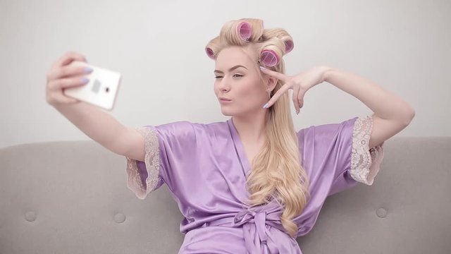 Happy cheerful blonde woman in purple bathrobe and curled hair using smartphone sitting on a couch, making selfies or video. Technology, free time, communication concept. Prores 422.