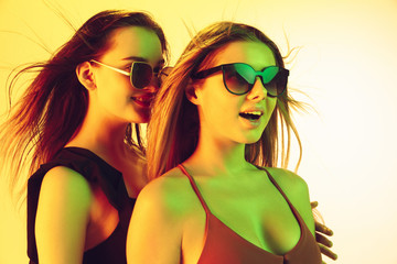 Beautiful young girls' portrait isolated on yellow studio background in neon light. Women in fashionable bodysuits. Facial expression, summer, weekend, beauty, resort concept. Vacations, youth.