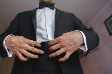 Groom in black tuxedo and bowtie correct his buttons on white shirt. Wedding. Details