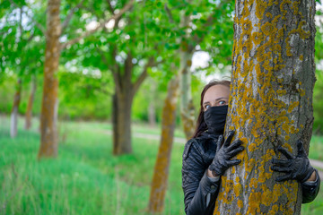Fototapeta premium Young woman in black medical mask and gloves hugging tree in woodland. Adult female enjoying nature in countryside in period coronavirus epidemic infection.