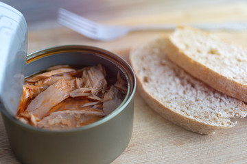 Close up of open canned tuna in red oil, fork and bread on wooden table, top view