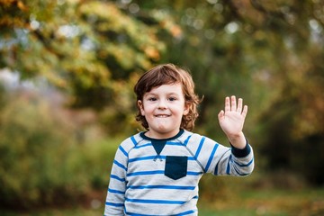 Summer, childhood, leisure, human gesture concept - happy little boy waving hand. Portrait of a little boy in the woods waving his hand. Portrait of a boy.