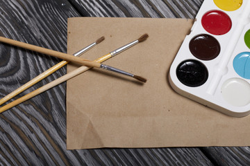 Watercolor for painting, brushes and a sheet of paper. They lie on pine boards painted with white and black paint.