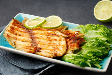 Grilled fish with fresh herbs and lime in a rectangular blue plate. Dark background.