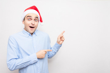 Guy in Santa Claus hat is surprised and points fingers towards, portrait, white background, copy...
