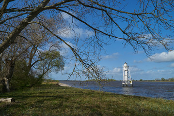 scenic white lighthouse on the island Elsflether Sand in the river Weser seen behind branches of a large willow tree on a bright spring day with vivid blue sky