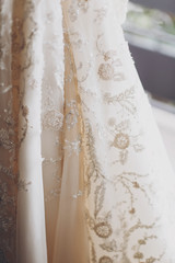 Stylish luxury beige wedding gown with lace floral pattern in light. Close up. Modern wedding dress hanging at window in soft morning light. Bridal morning preparations and boudoir