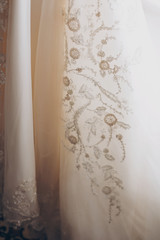 Stylish luxury beige wedding gown with lace floral pattern in light. Close up. Modern wedding dress hanging at window in soft morning light. Bridal morning preparations and boudoir