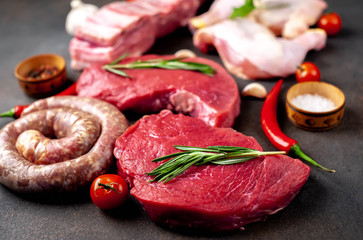 Various types of grill and barbecue. Raw beef steaks, sausages, pork ribs, chicken legs with spices on a stone background