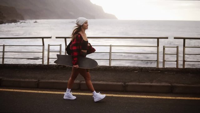Side view of muscular, sporty woman in hat, coat and shorts enjoying time by the seaside on a cloudy day at sunset, walking freerly, holding a skateboard. Wind fluttering her hair. Stormy ocean on