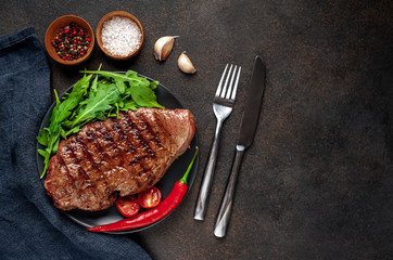 Grilled beef steak with spices and herbs on a black plate with cutlery on a stone background with copy space for your text