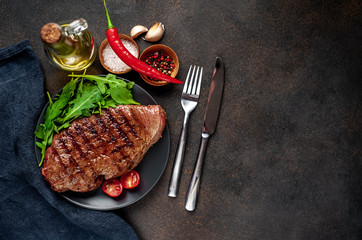Grilled beef steak with spices and herbs on a black plate with cutlery on a stone background with copy space for your text