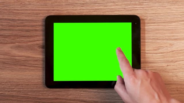 Top view of male hands using tablet with green screen on wooden desk. His fingers touching and swiping on screen.
