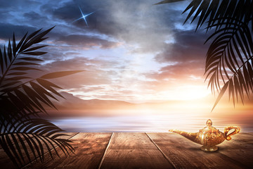 Aladin's lamp on the background of a sea evening landscape with sunset. Palm tree branches, silhouettes, sunlight. Wooden table. Night view, open-air seascape. 3D illustration