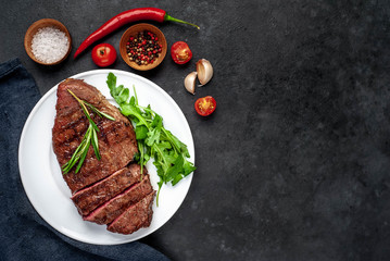 Fototapeta na wymiar Grilled beef steak with spices, tomatoes and herbs on a white plate on a stone background with copy space for your text.