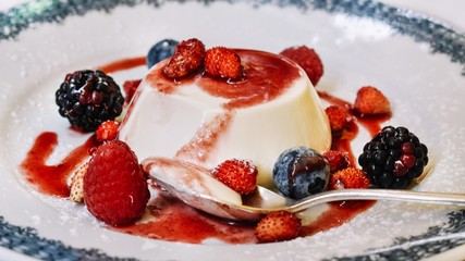 Side view of a plate of cooked cream flan (panna cotta) garnished with strawberry sauce and fresh red berries sprinkled with icing sugar