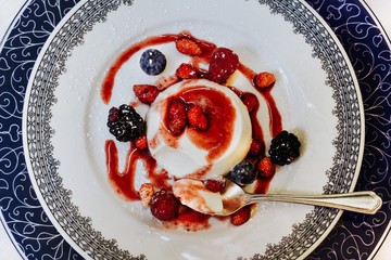 Top view of a plate of cooked cream flan (panna cotta) garnished with strawberry sauce and fresh red berries sprinkled with icing sugar
