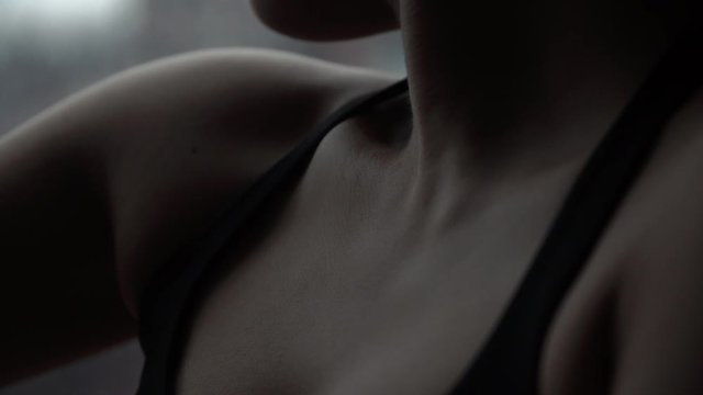 Soft skin and chest of woman in fitness attire sitting by window in a modern dance studio in silhouette, close up tilt down from nick down