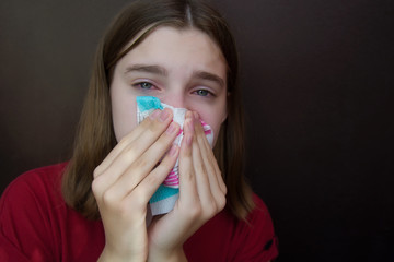A young girl with an allergy and with red eyes blows her nose in a napkin. Dark background....