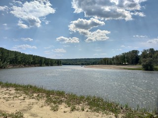 Shore over the gray river. Blue sky with white clouds. Green forest on the shore.