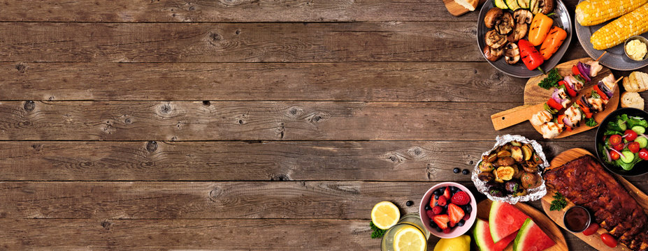 Summer BBQ or picnic food corner border over a rustic wood banner background. Assorted grilled meats, vegetables, fruits, salad and potatoes. Overhead view with copy space.