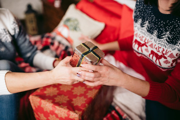 Two young woman in Christmas sweaters exchange gifts sitting on the bed with lots of pillows