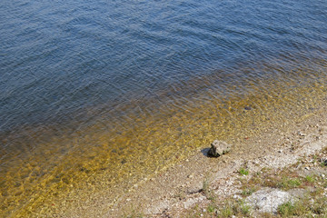 Coastline washing by waves of river, sunlight playing in calm waters & changing water color to gold tones