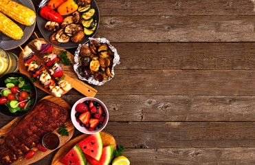 Summer BBQ or picnic food side border over a rustic wood background. Grilled meats, vegetables, fruits, salad and potatoes. Top view with copy space.