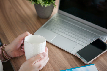 female hands holding a white cup, on the background of a computer and a phone