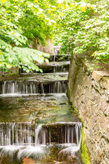 Stepped cascading waterfall in the greenery of bushes and trees with stone walls along. Summer. Vertical orientation.
